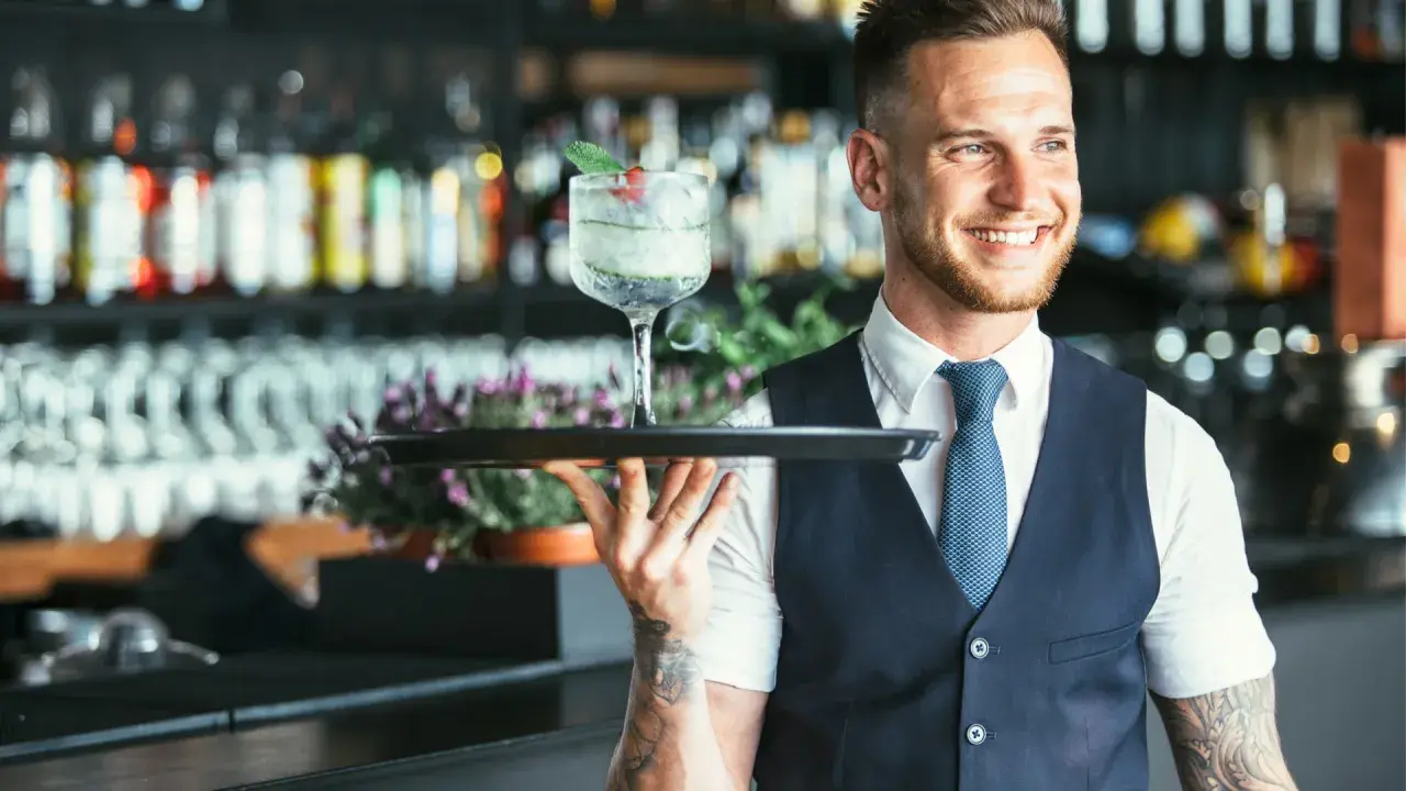 Lächelnder Barkeeper mit Tätowierungen hält ein Tablett mit einem erfrischenden Drink. Er serviert von der rechten Seite.