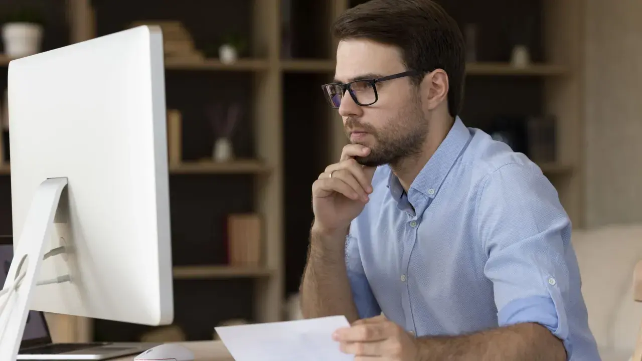 Mann mit Brille am Schreibtisch, der nachdenklich auf den Bildschirm blickt, um alle Mails in Outlook anzuzeigen.