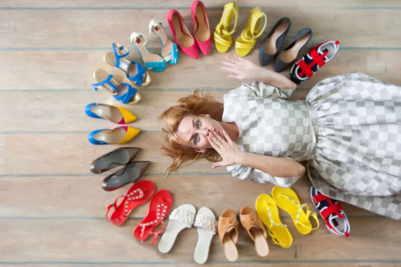Frau liegt inmitten einer bunten Schuhsammlung, die sie gerade aussortiert. Von High Heels bis Sneakern ist alles dabei.