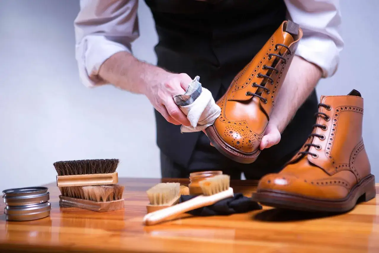 Mann poliert braune Lederschuhe mit einem Tuch. Zubehör wie Bürsten und Schuhcreme liegen bereit für die Lederpflege.