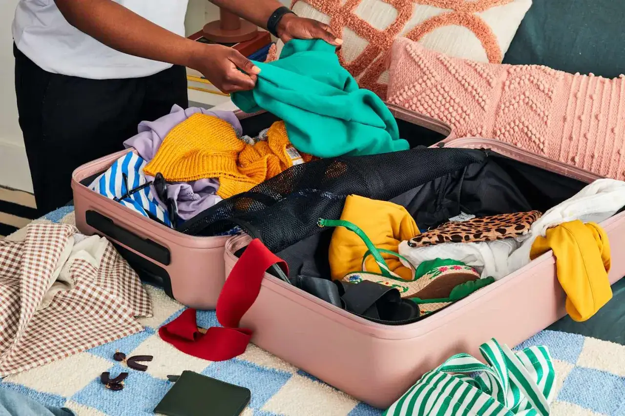 Person packt eine Reisetasche mit bunter Kleidung, Schuhen und Accessoires.