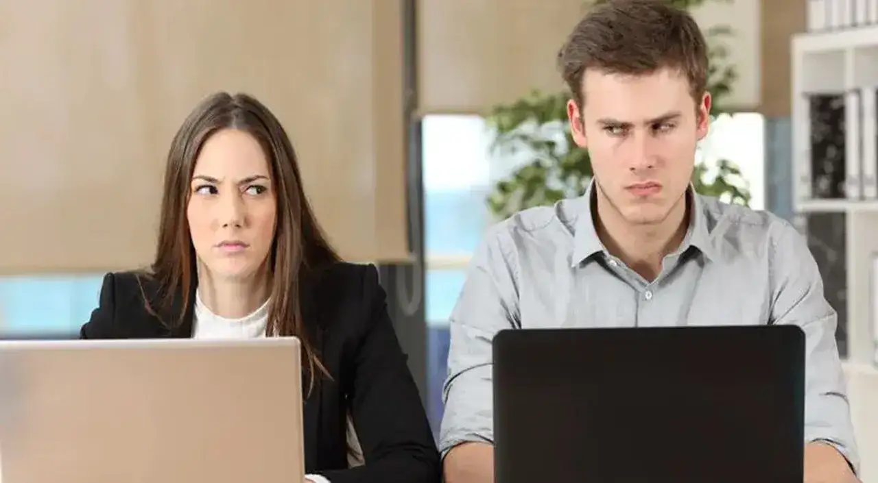 Zwei junge, neidische Kollegen blicken sich misstrauisch an, w&auml;hrend sie an ihren Laptops arbeiten.