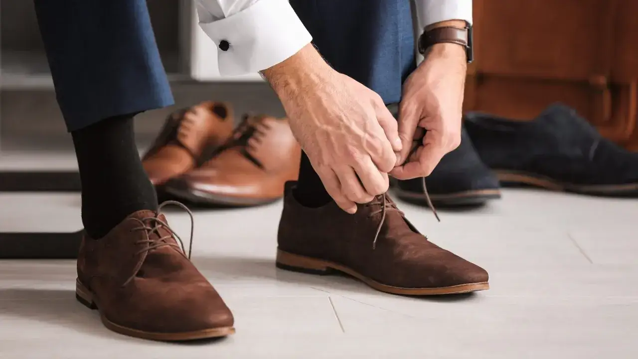 Mann schn&uuml;rt braune Wildlederschuhe, die perfekt zu seinem Anzug passen. Mehrere andere Anzugschuhe liegen bereit.