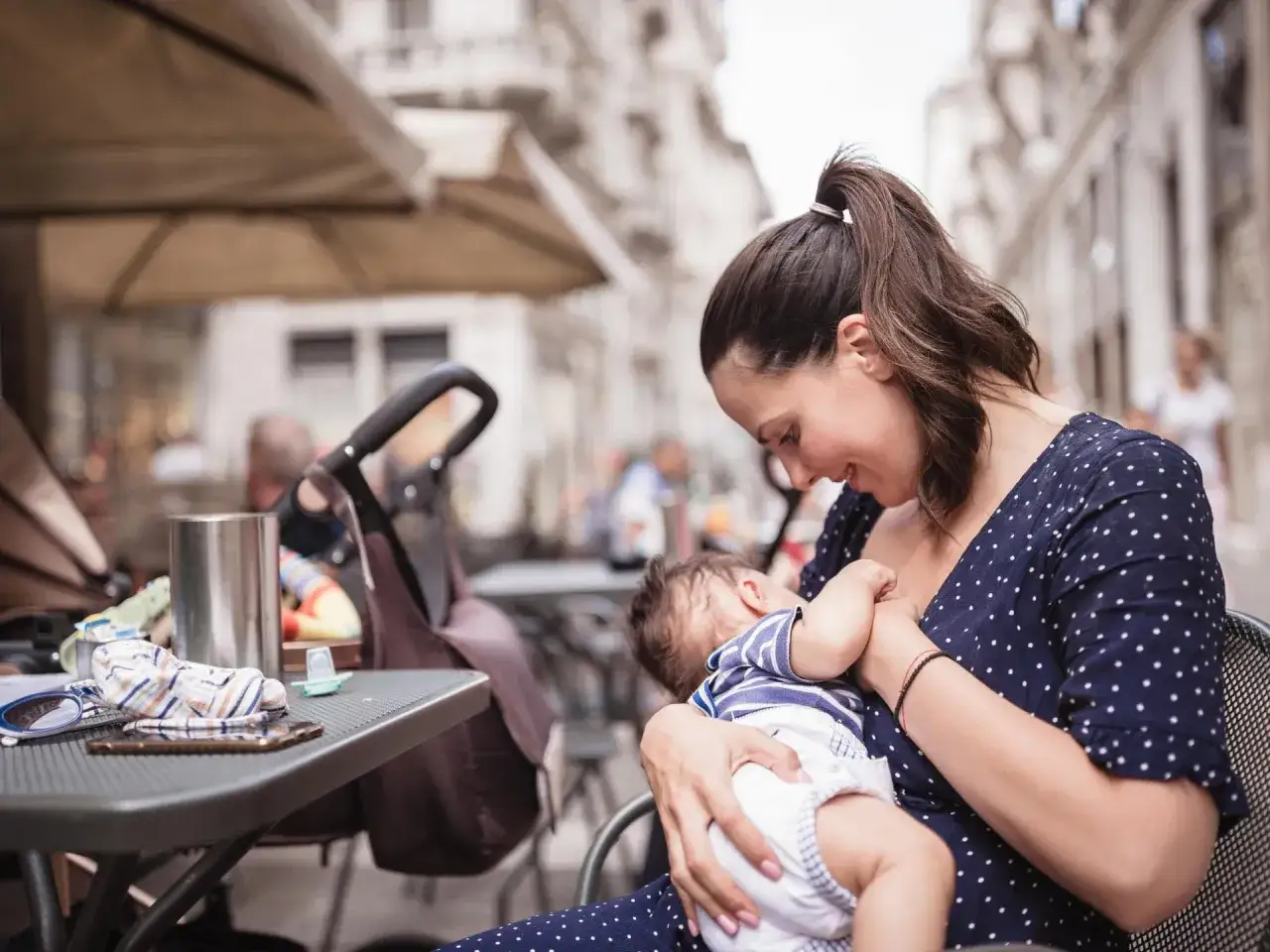 Eine Mutter stillt ihr Baby im Freien an einem Tisch. Ein Kinderwagen steht daneben.