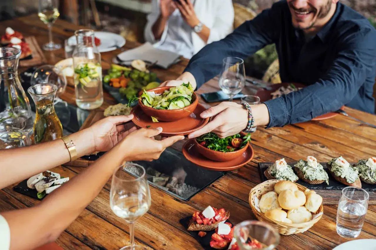 Ein fr&ouml;hlicher restaurant besuch mit Freunden. Sie teilen sich Salate und Bruschetta an einem Holztisch, dazu gibt es Wein und Wasser.