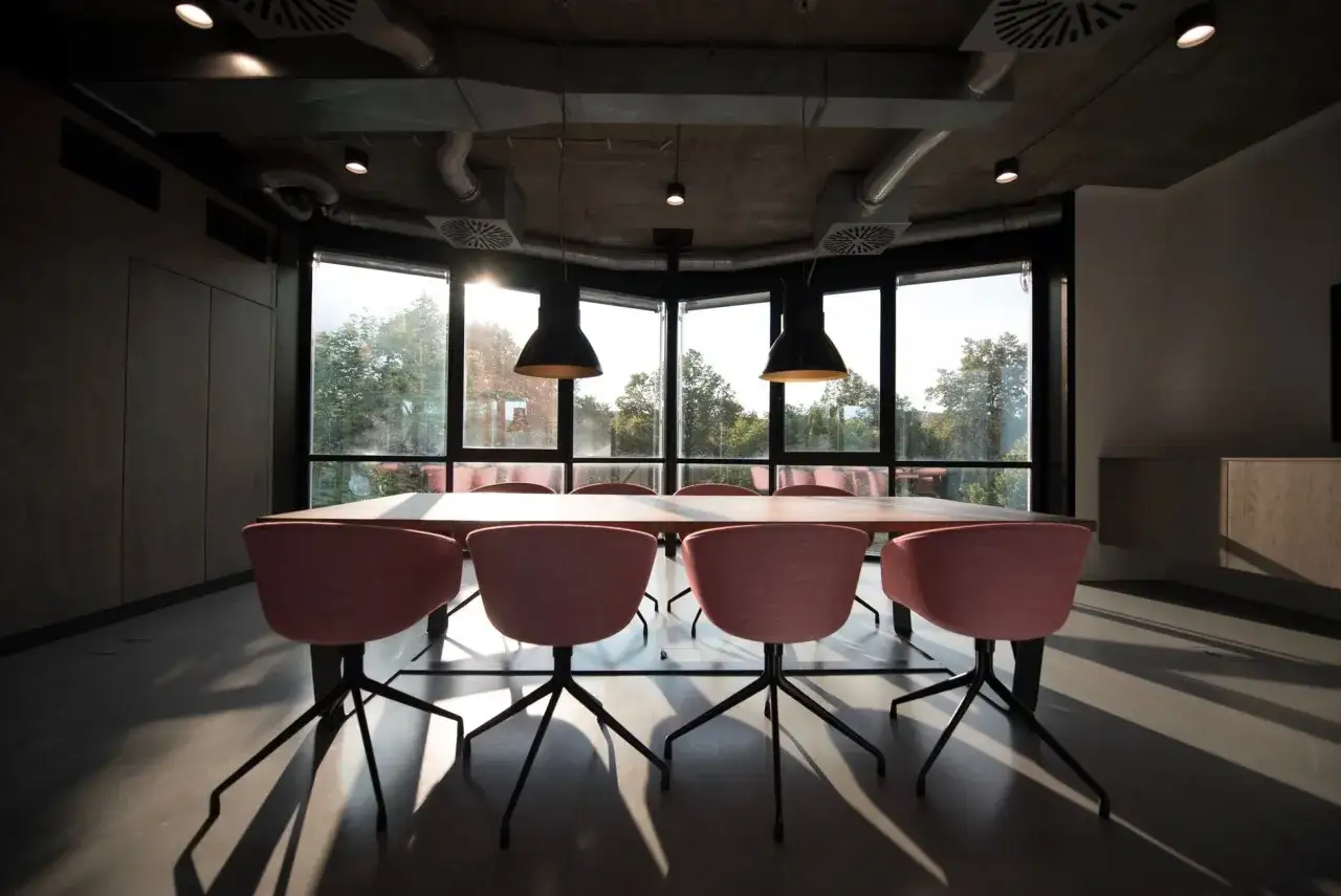 Moderne Besprechungsraum mit U-förmiger Sitzordnung. Rosa Stühle um einen Holztisch, große Fenster mit Blick ins Grüne.