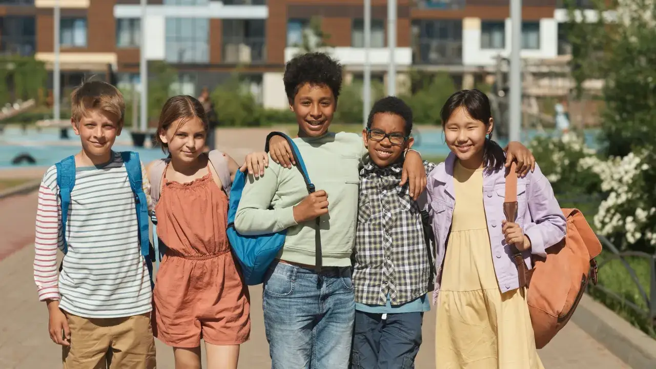 Fünf Kinder mit Rucksäcken, die freundlich in die Kamera lächeln. Sie zeigen, wie gute Benimmregeln bei Jugendlichen aussehen können: Respekt und Freundschaft.