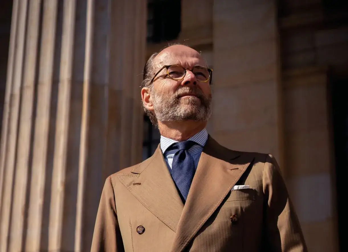 Bernhard Roetzel, ein Mann mit Bart und Brille, trägt einen braunen Anzug und eine blaue Krawatte vor einer Säule.