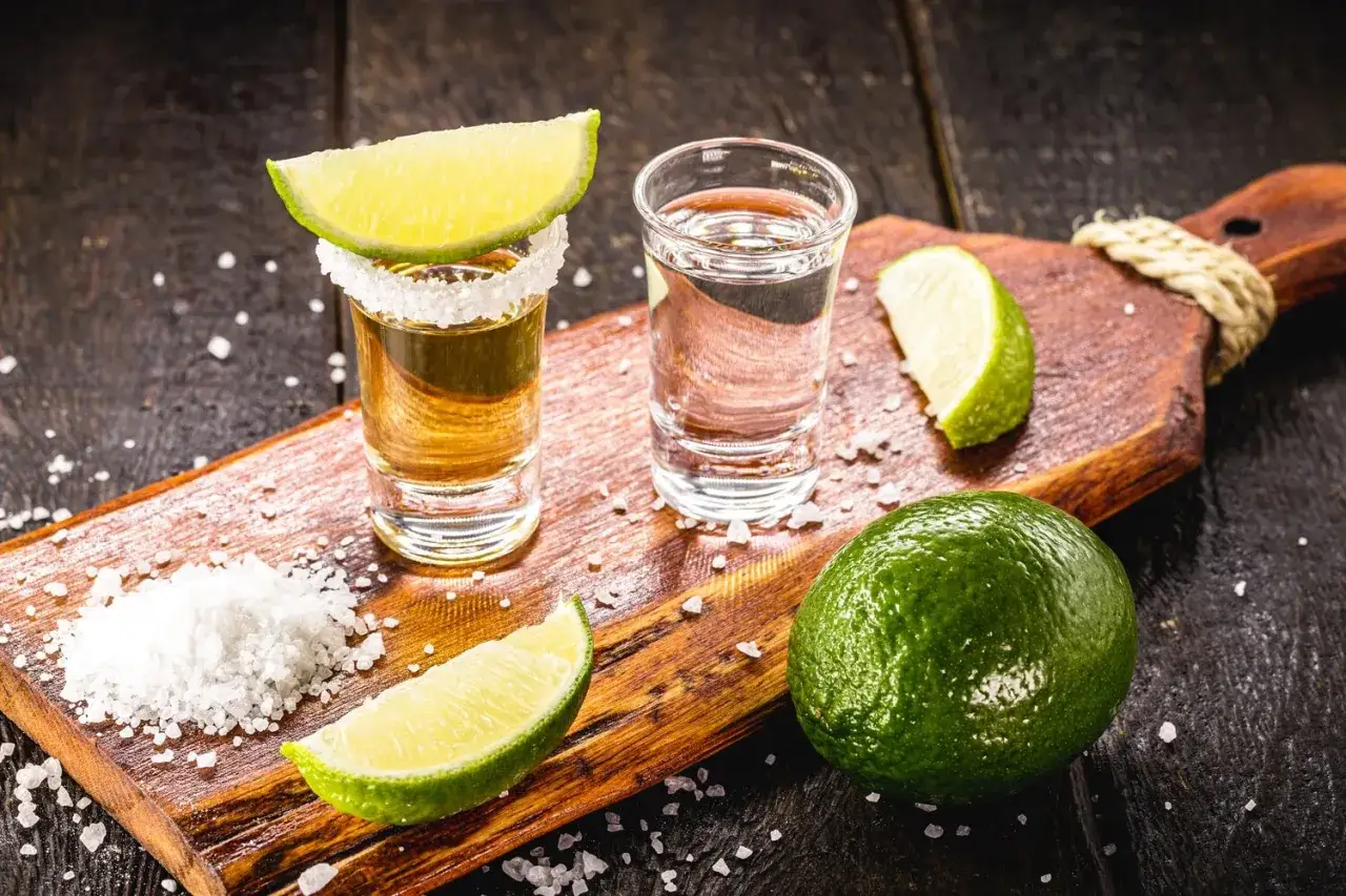 Zwei Tequila-Gläser, Salz, Limettenspalten und eine ganze Limette auf einem Holzbrett – so kann man Tequila richtig trinken.