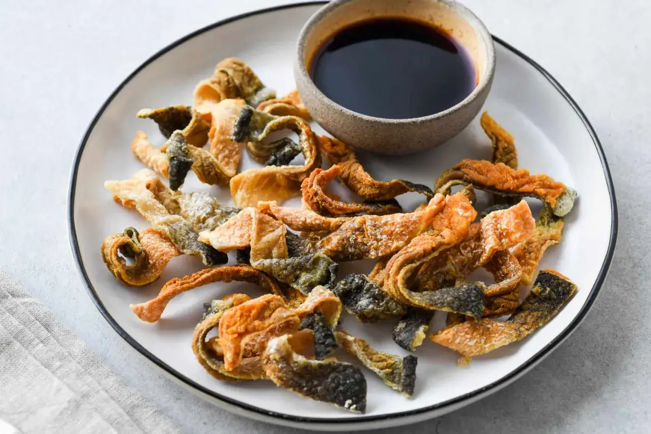 Knusprige, frittierte Fischhaut-Chips liegen auf einem Teller, daneben eine Schale mit dunkler So&szlig;e. Ein ungew&ouml;hnlicher Snack zum Fischhaut essen.