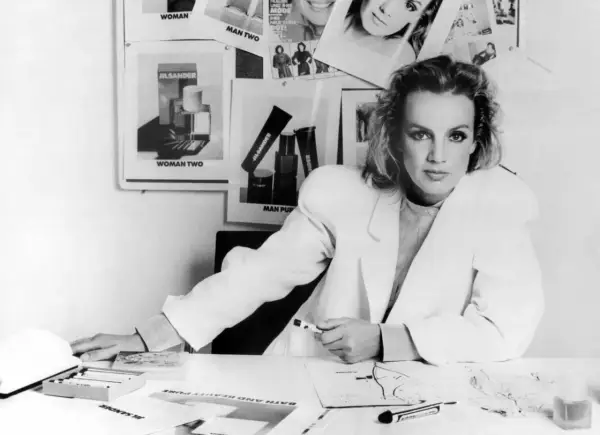 Jil Sander Designerin am Schreibtisch, umgeben von Entwürfen und Produktbildern ihrer Marke.